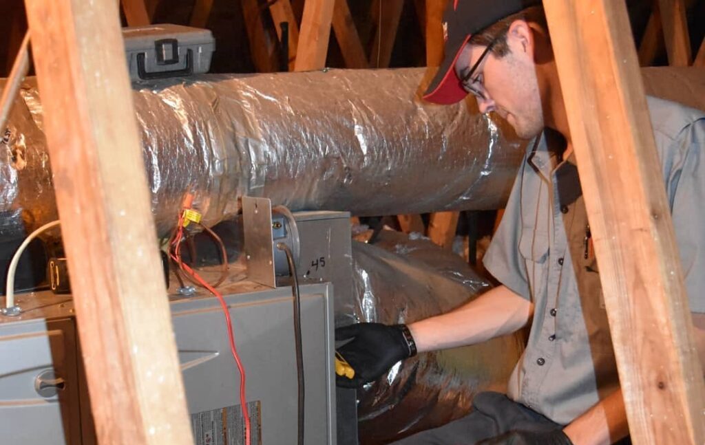 Male technician kneeling in an attic, wearing gloves and a cap, working on a furnace with exposed ductwork and wiring nearby.