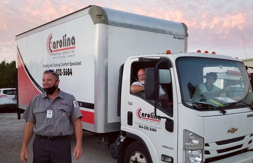 White delivery truck with Carolin Heating Service Inc logo pulling a trailer; a technician in gray uniform stands outside smiling next to the cab.