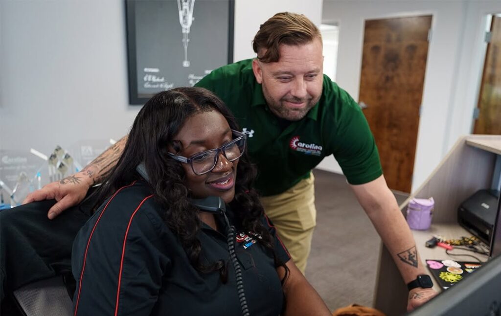 Woman with headset seated at a computer as a man in a green polo leans in to help her in an office setting.