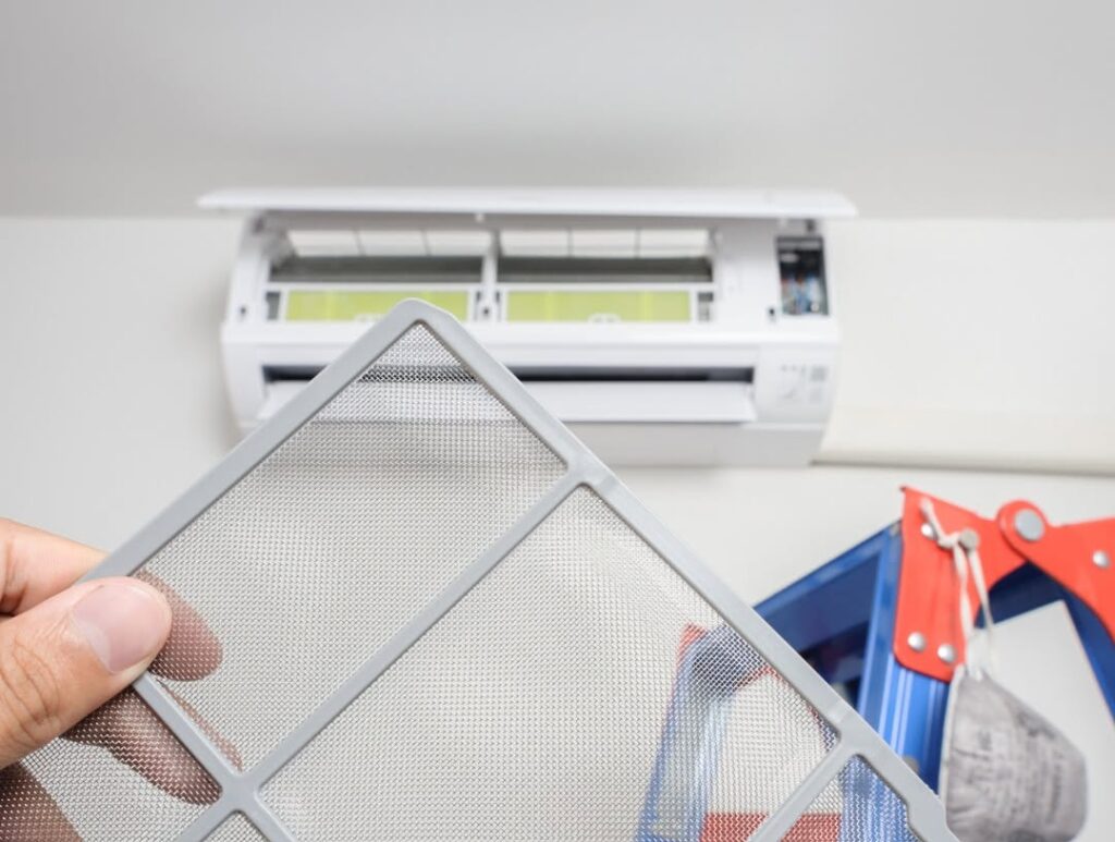 Person holding a metal mesh air filter in front of a wall-mounted split-system air conditioner unit, mid-replacement.