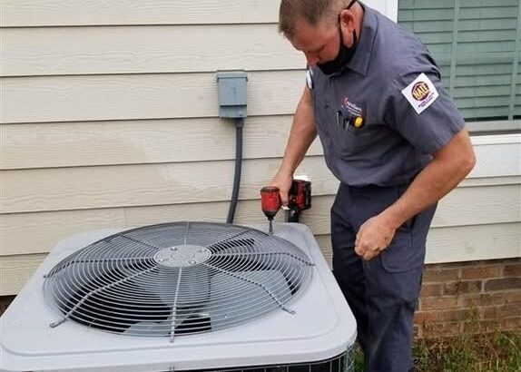 HVAC technician wearing a mask services an outdoor air conditioner condenser with a tool beside a house window.