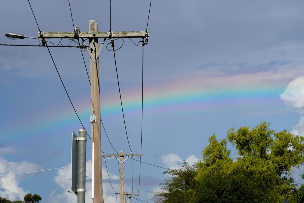Wood utility poles and power lines stretch across a blue sky, with a faint rainbow arcing in the background and trees on the right.